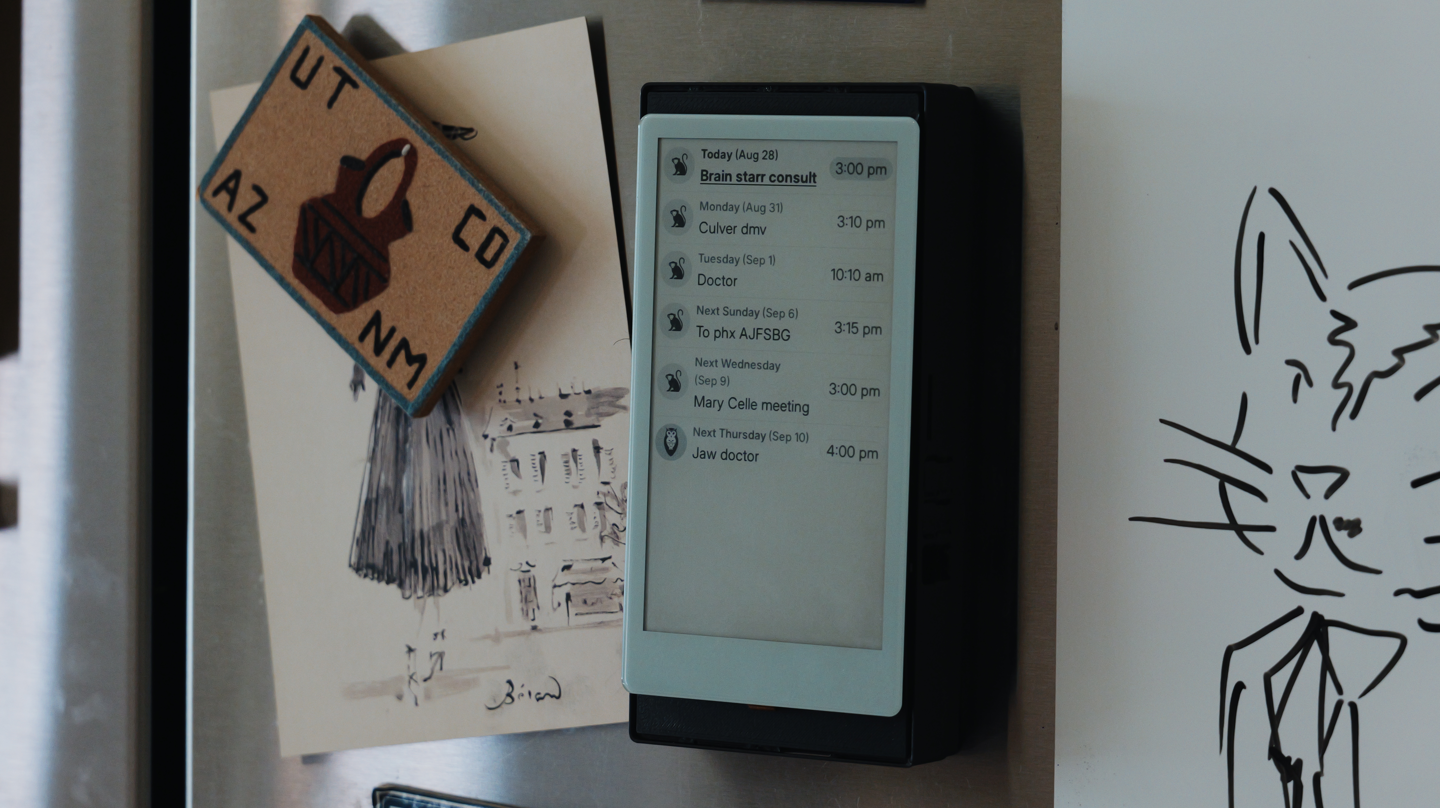A tall e-ink calendar mounted on a refrigerator beside postcards and a cat drawing