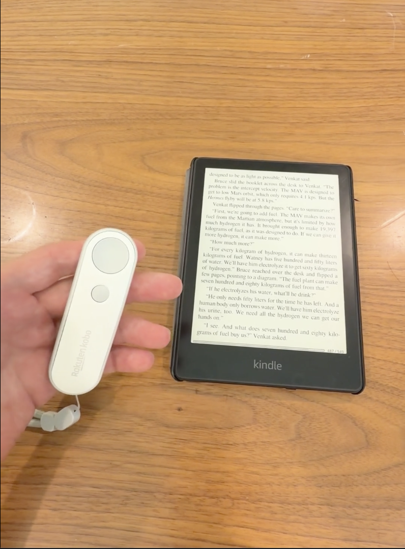 A hand holding a white Bluetooth page-turning remote beside a Kindle on a wooden table